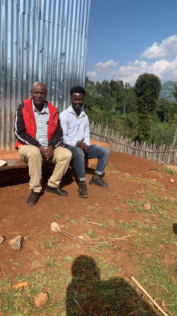 Two men sitting on a wooden bench outdoors with a scenic background