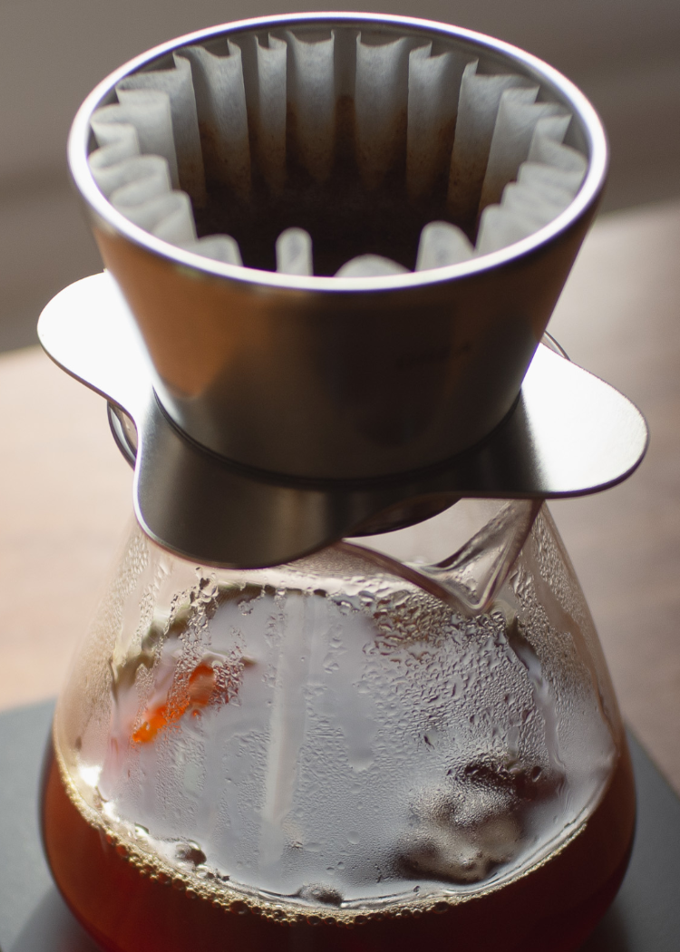 A pour-over coffee setup with a metal cone filter resting on a glass carafe filled with dark coffee, photographed close up on a scale.