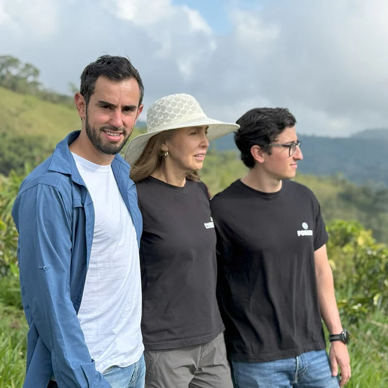Three people standing outdoors with a scenic background of green hills and mountains.