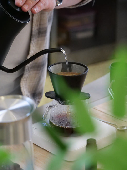 A person pouring water from a kettle into a coffee maker, which is placed on a kitchen scale with ground coffee inside.