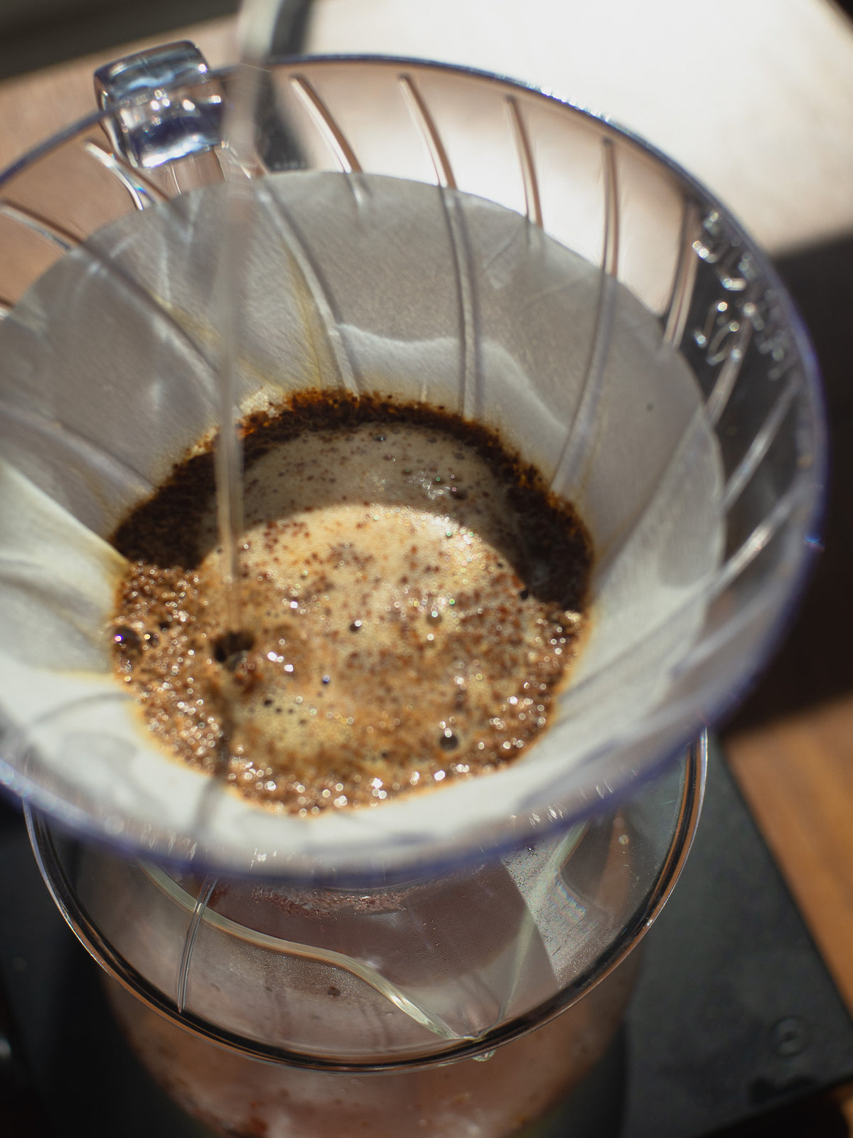 A close-up of a pour-over coffee setup with dark coffee grounds filtering through a white paper cone into a clear glass carafe.