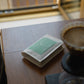 A small white coffee bag with a mint green label reading 'Abner & Vanessa' sits on a wooden table next to a pour-over coffee maker and a glass carafe.