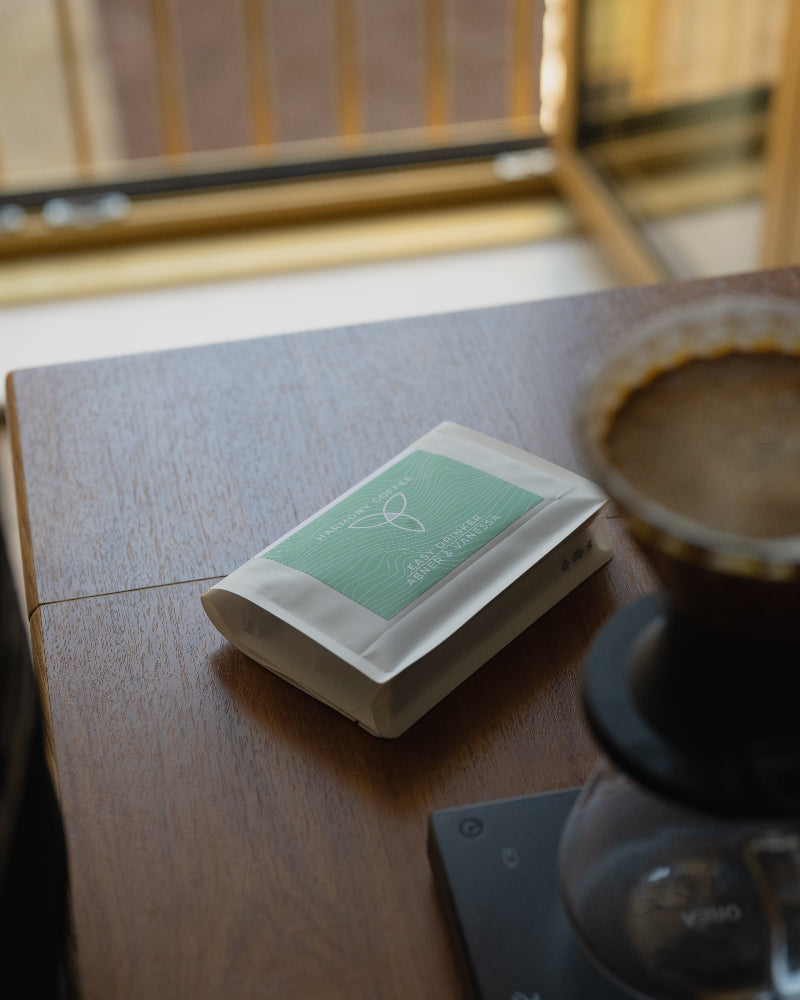 A small white coffee bag with a mint green label reading 'Abner & Vanessa' sits on a wooden table next to a pour-over coffee maker and a glass carafe.