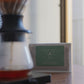 A bag of Harmony Coffee 'Easy Drinkers Abner & Vanessa' coffee sits on a wooden table next to a glass pour-over coffee maker with water dripping into it.