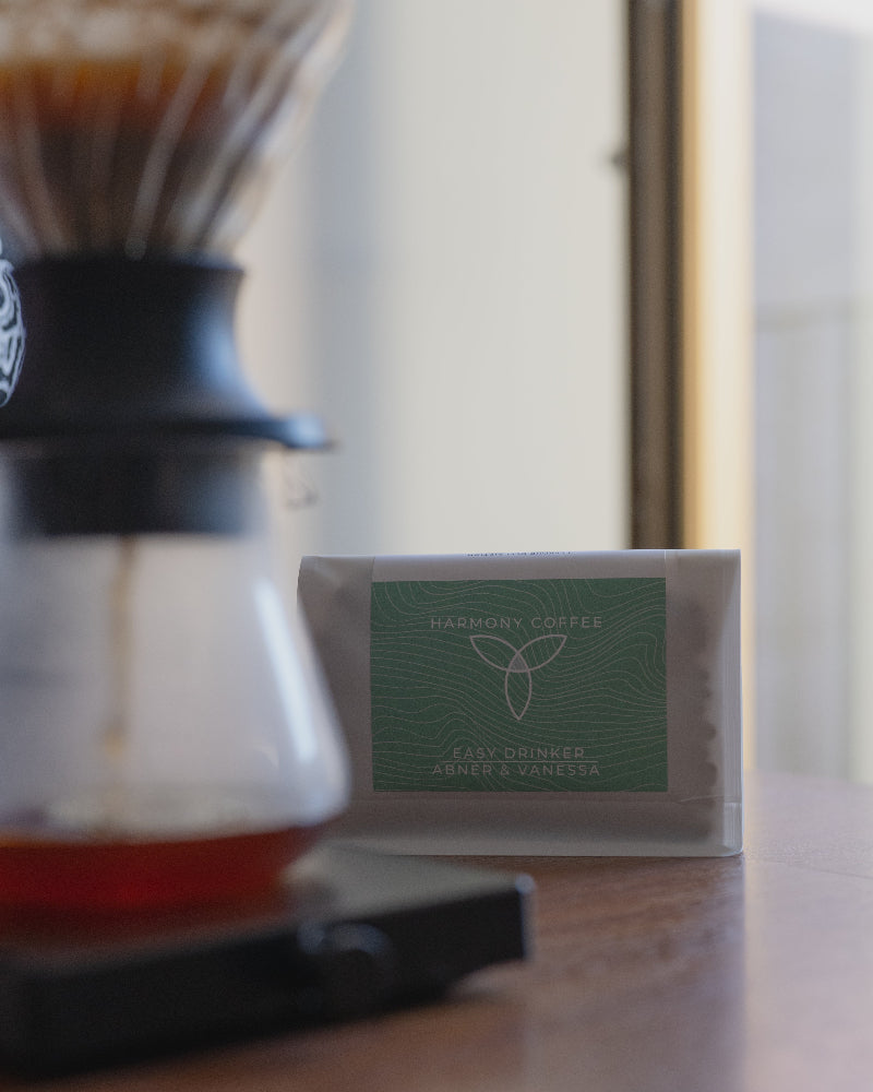 A bag of Harmony Coffee 'Easy Drinkers Abner & Vanessa' coffee sits on a wooden table next to a glass pour-over coffee maker with water dripping into it.