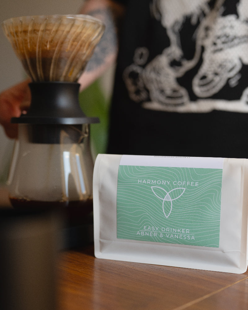 A bag of Harmony Coffee 'Easy Drinker Abner & Vanessa' coffee beans sits on a wooden table next to a glass pour-over coffee maker with brewed coffee.