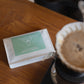 A white pouch of Harmony Coffee Easy Drinker Abner & Vanessa coffee sits on a wooden table next to a pour-over coffee maker with brewed coffee in a paper filter.