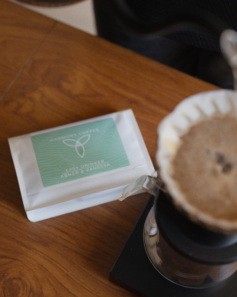 A white pouch of Harmony Coffee Easy Drinker Abner & Vanessa coffee sits on a wooden table next to a pour-over coffee maker with brewed coffee in a paper filter.