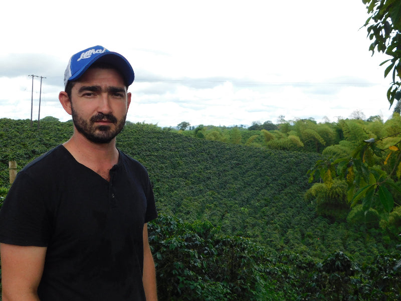 Arturo Arango wearing a blue cap and black shirt standing in front of a El Paraiso Coffee Farm. 