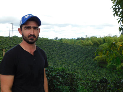 Arturo Arango wearing a blue cap and black shirt standing in front of a El Paraiso Coffee Farm. 