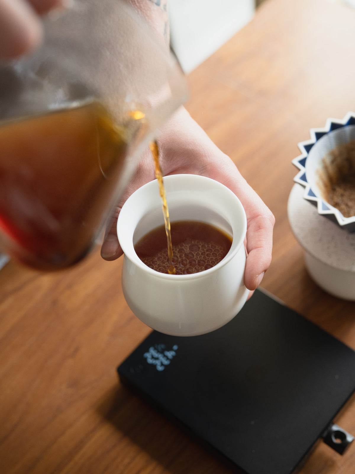 A hand pouring brewed coffee from a glass carafe into a small white cup on a wooden table, with a black scale and a coffee filter visible in the background.