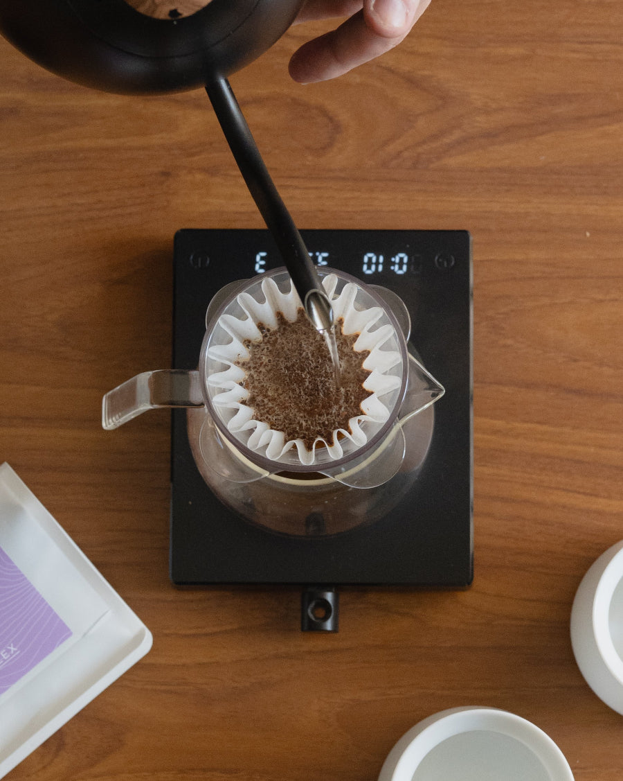 A top-down view of a person pouring hot water from a black kettle into a glass pour-over filter on a digital scale, set on a wooden table. 