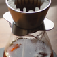 A pour-over coffee setup with a metal cone filter resting on a glass carafe filled with dark coffee, photographed close up on a scale.