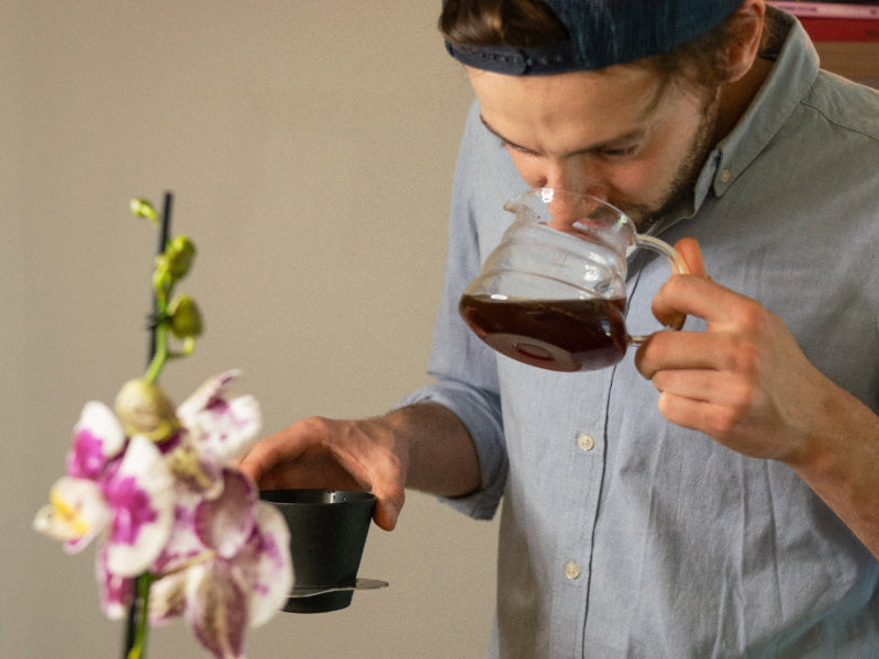 Man holding a glass carafe of coffee with a flower arrangement in the foreground