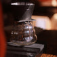 Clear glass coffee brewing device with a black set of scales on a wooden surface.