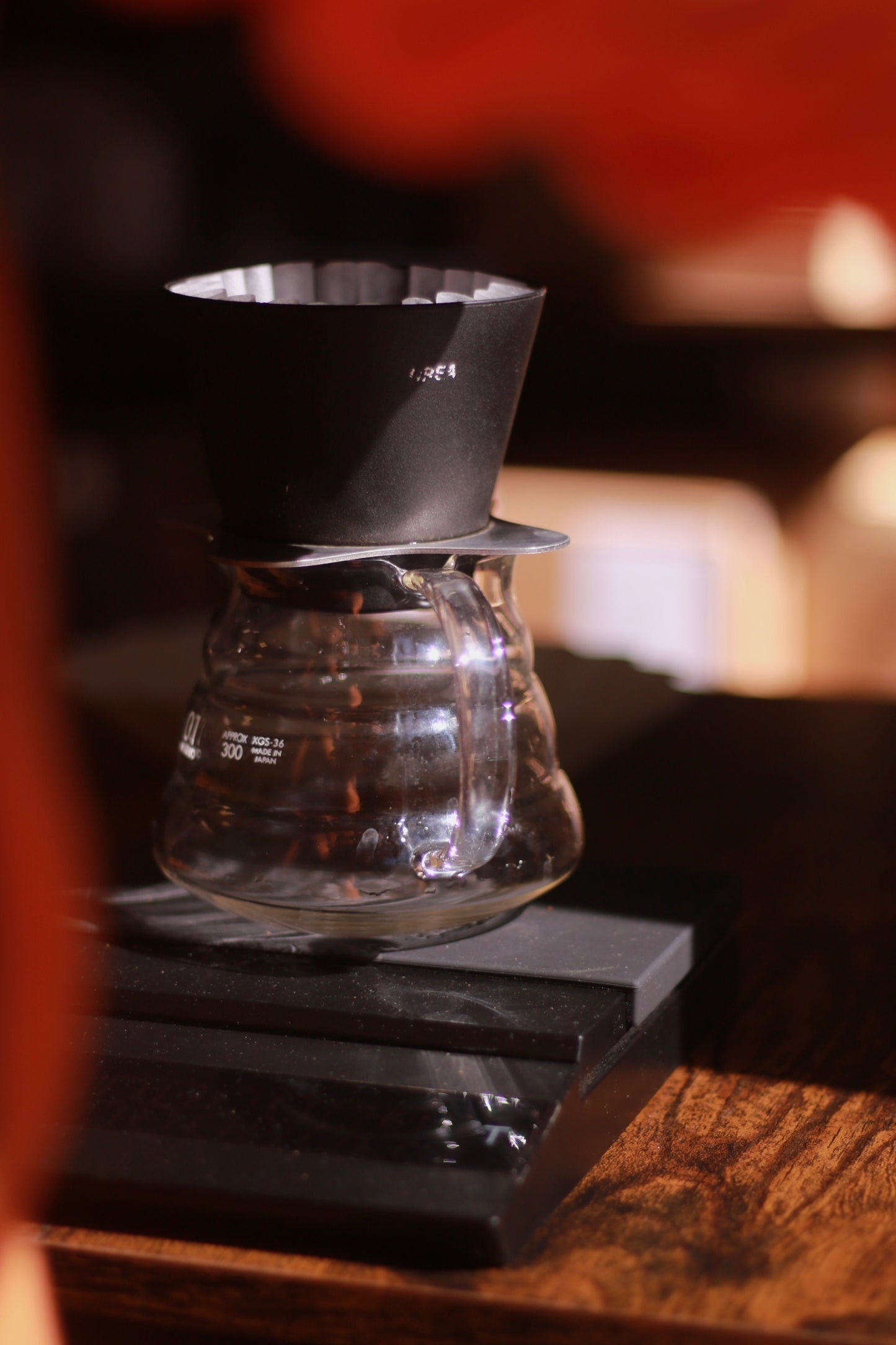 Clear glass coffee brewing device with a black set of scales on a wooden surface.