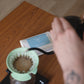 A close-up lifestyle shot of a person pouring hot water from a black kettle into a mint green cone filter pour-over coffee maker placed on a digital scale, with a coffee bag on a wooden table.