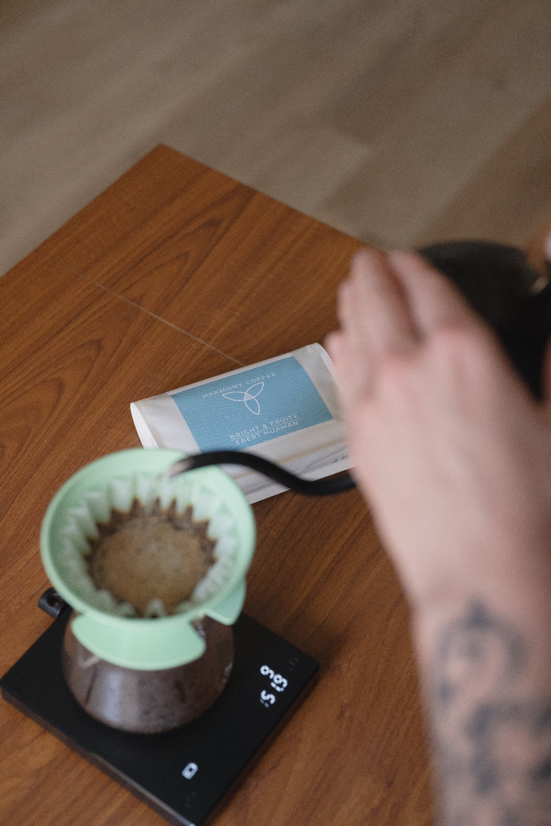 A close-up lifestyle shot of a person pouring hot water from a black kettle into a mint green cone filter pour-over coffee maker placed on a digital scale, with a coffee bag on a wooden table.