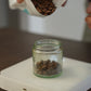 Person pouring coffee beans from a Harmony coffee bag into a glass jar on a white set of Acaia scales.