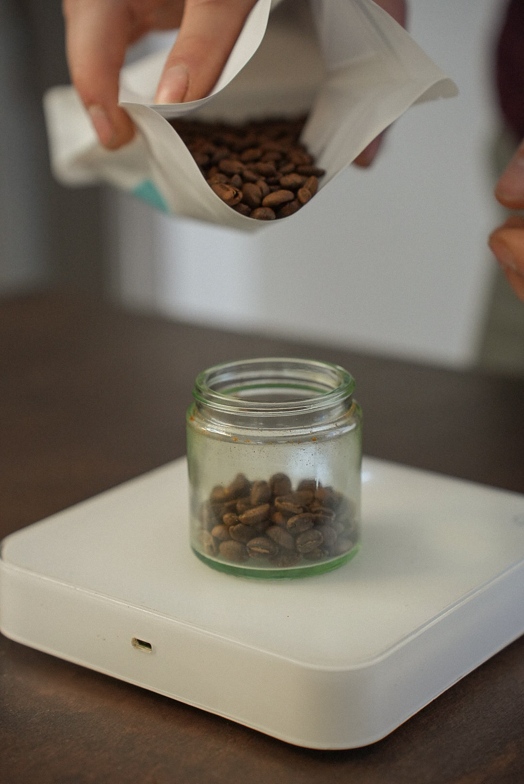Person pouring coffee beans from a Harmony coffee bag into a glass jar on a white set of Acaia scales.
