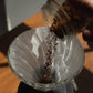 A close-up of coffee grounds being poured from a glass jar into a white paper cone filter over a glass pour-over dripper on a wooden table.
