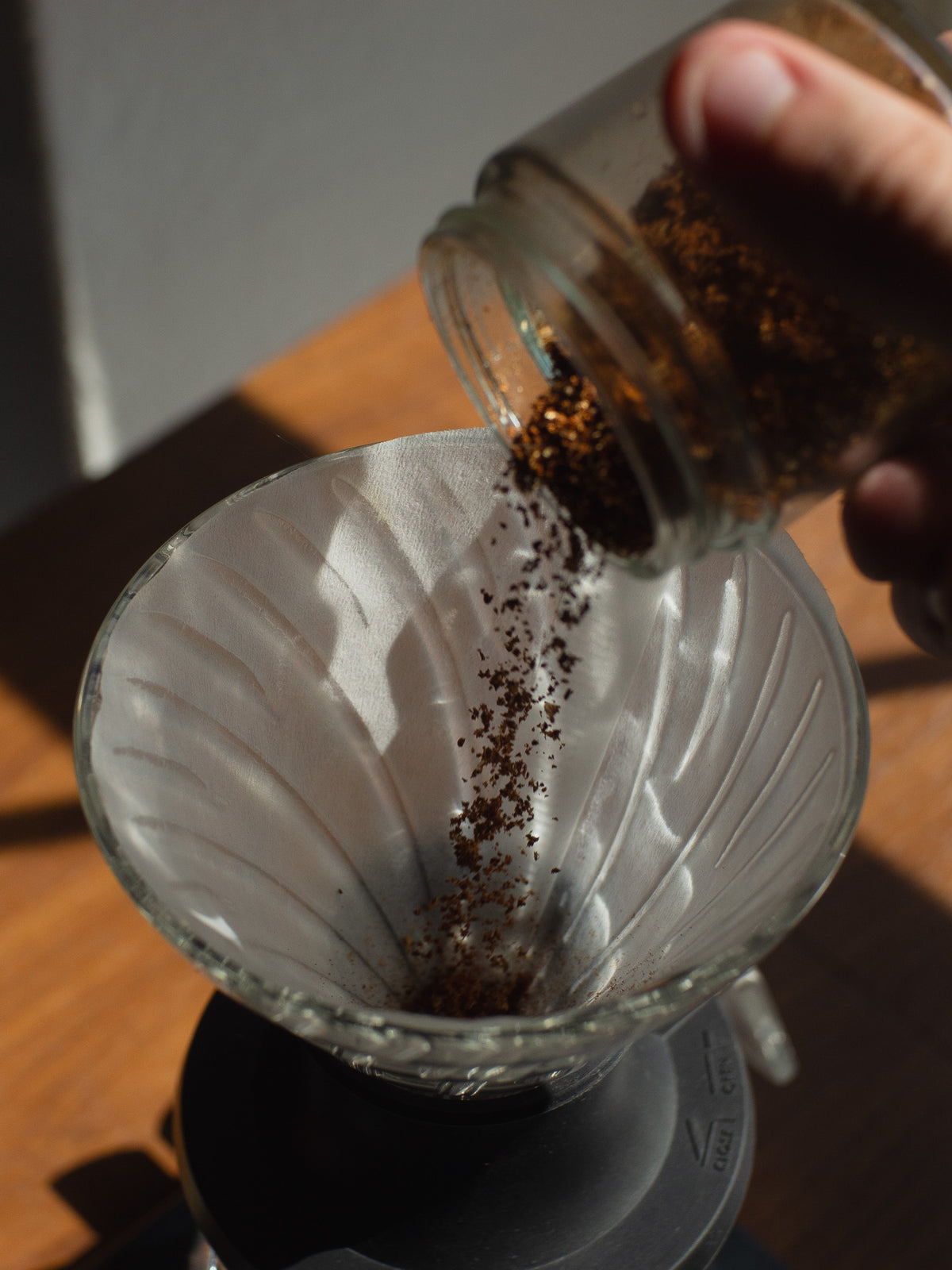 A close-up of coffee grounds being poured from a glass jar into a white paper cone filter over a glass pour-over dripper on a wooden table.