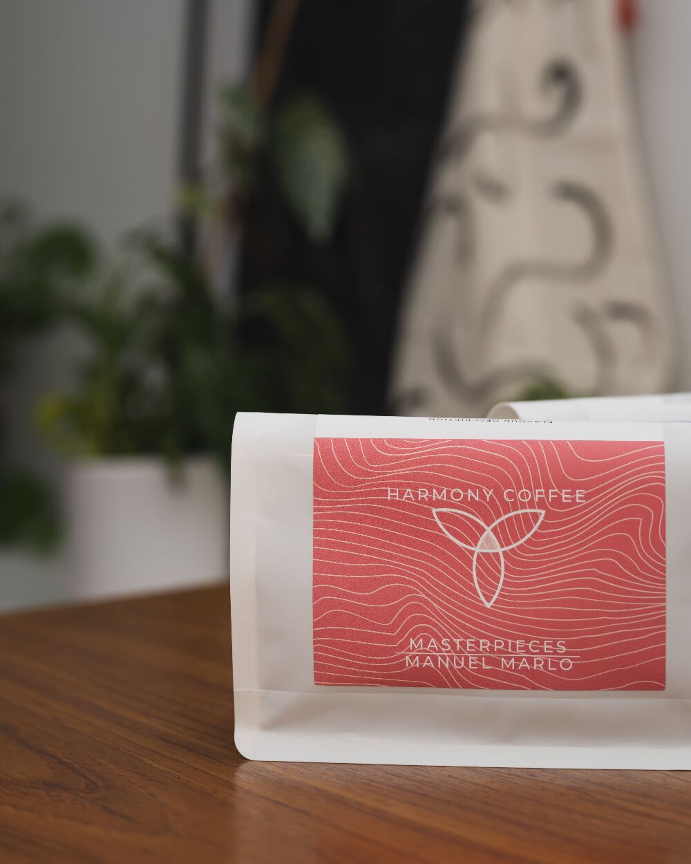 A bag of Harmony Coffee Masterpieces Manuel Marlo Gesha on a wooden table, with a coral label and white wavy line art
