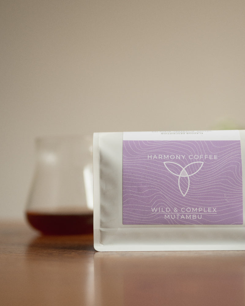 A stand-up coffee bag labeled 'Harmony Coffee' and 'Wild & Complex Mutambu' sits on a wooden surface with a blurred glass of coffee in the background against a warm beige wall.