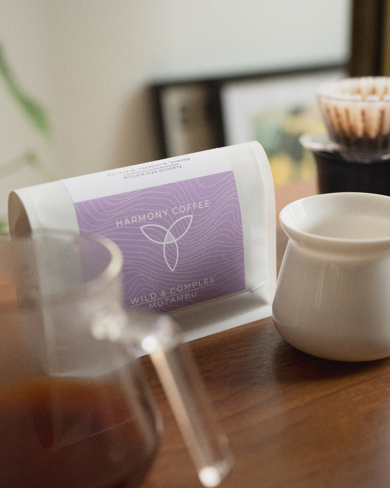 A bag of Harmony Coffee 'Wild & Complex Mutambu' coffee beans sits on a wooden table next to a white ceramic cup, with a glass carafe of brewed coffee in the foreground and a hand-drip coffee maker blurred in the background.