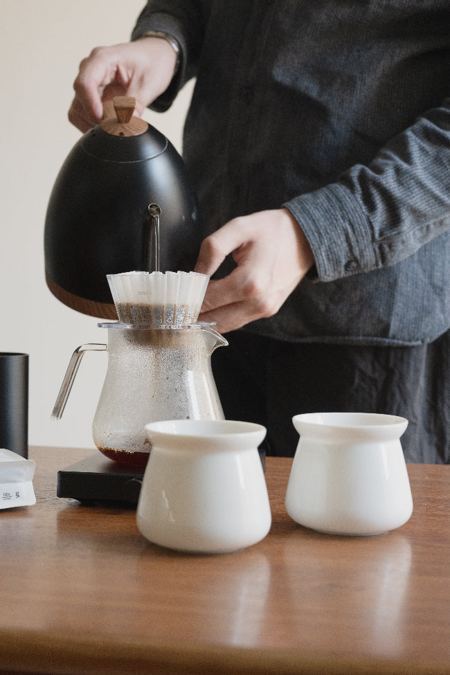 A person in a dark gray shirt pour-runs coffee from a matte black kettle into a glass carafe with a white paper filter, with two small white ceramic cups on a wooden table in the foreground.