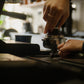 Person using a coffee tamper to manually tamp a bed of coffee in a cafe setting