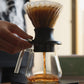 A hand holds the side of a glass pour-over coffee maker with a black filter base, showing coffee dripping into a glass carafe on a scale in a warm indoor setting.