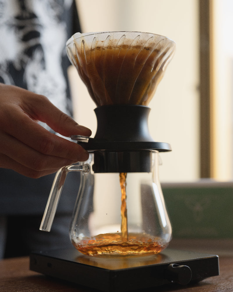 A hand holds the side of a glass pour-over coffee maker with a black filter base, showing coffee dripping into a glass carafe on a scale in a warm indoor setting.