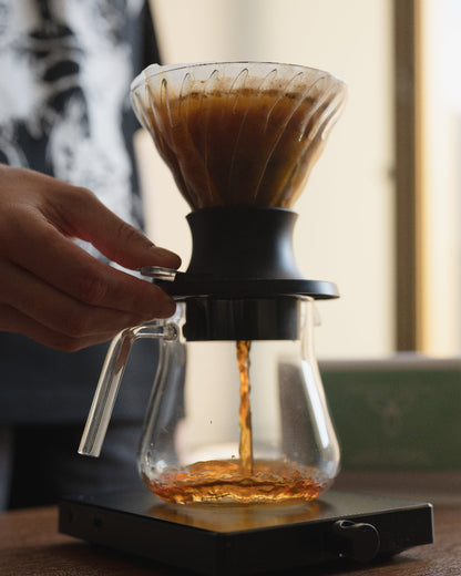 A hand holds the side of a glass pour-over coffee maker with a black filter base, showing coffee dripping into a glass carafe on a scale in a warm indoor setting.