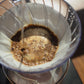 A close-up of a pour-over coffee setup with dark coffee grounds filtering through a white paper cone into a clear glass carafe.